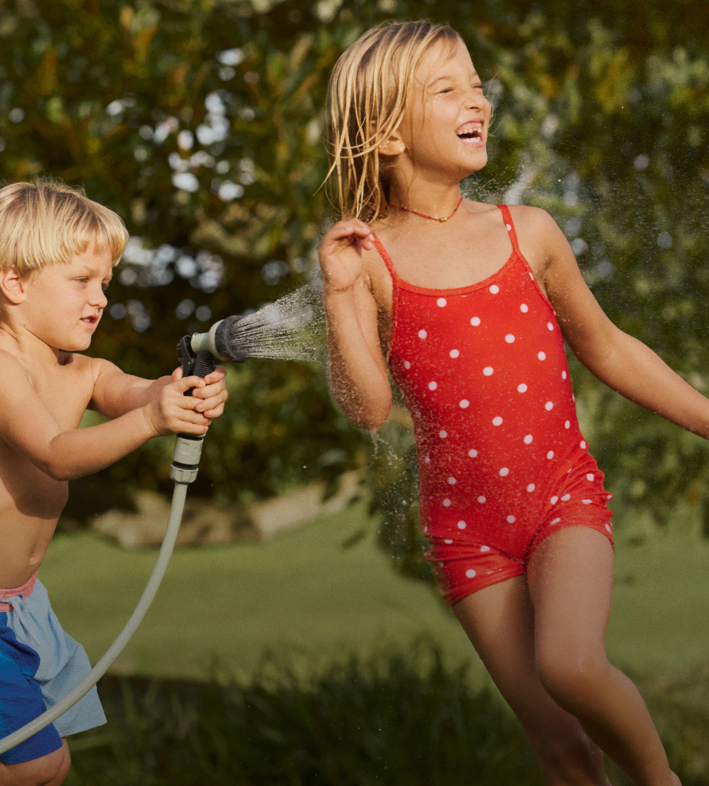 Two young children playing with a garden hose outdoors. Girl wearing red polka dot swimsuit, boy in blue shorts. Pixelated parrot graphic overlay