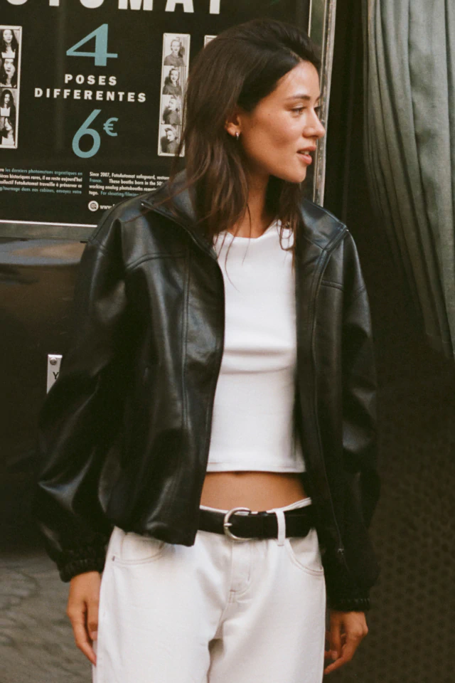 Woman wearing black leather jacket and white crop top with white pants and black belt, standing in front of photo booth signage