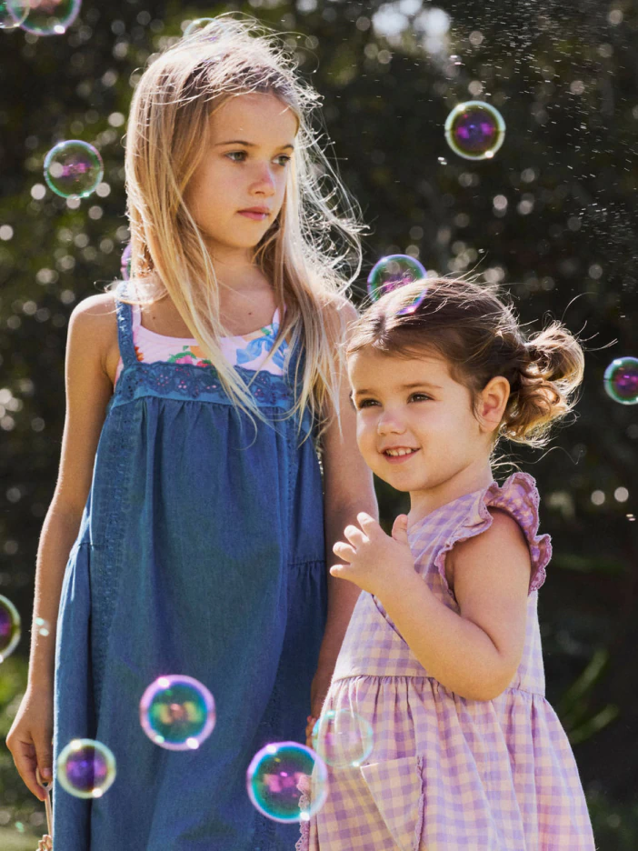Two young girls in summer dresses surrounded by soap bubbles. Older girl wears blue dress, younger girl wears lavender checkered dress