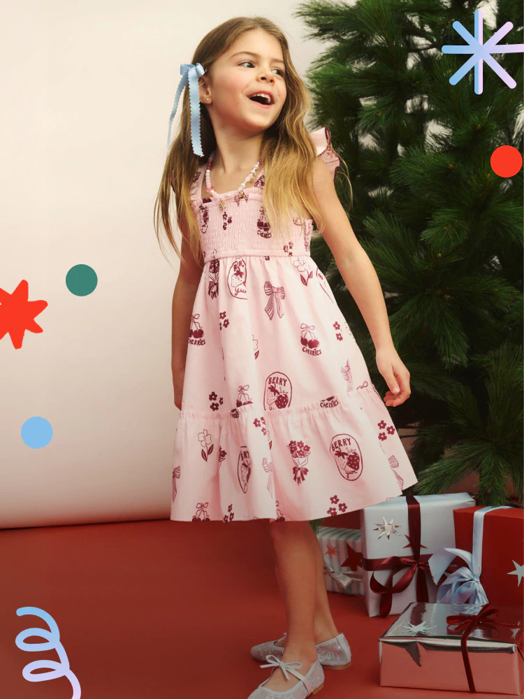 Young girl wearing pink dress with red berry print and blue ribbon bow, standing beside wrapped gifts and decorated Christmas tree
