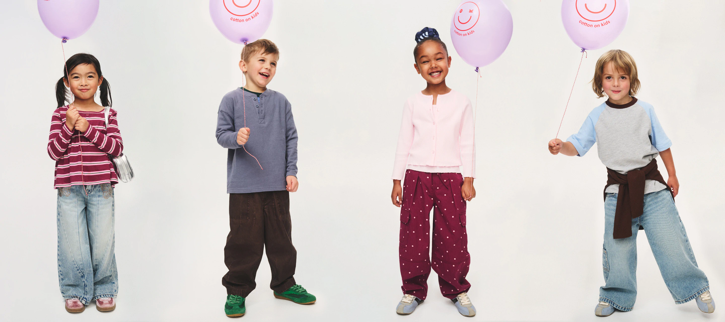 Four smiling children holding purple balloons with smiley faces, wearing casual striped and solid colored tops with jeans and sneakers