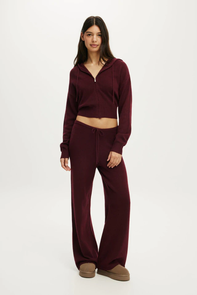 Woman wearing burgundy cropped zip-up hoodie and matching wide-leg sweatpants with beige slippers against neutral background