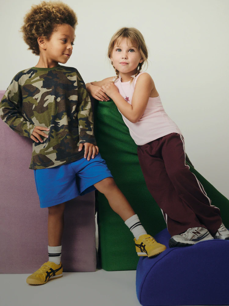 Two children posing together wearing colorful casual clothing including camouflage shirt, pink tank top, blue and maroon shorts, and yellow sneakers