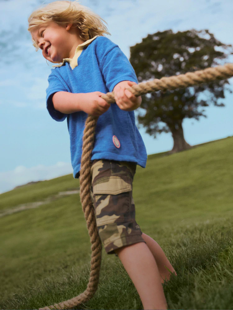 Young child in blue polo shirt and camouflage shorts holding a rope in a grassy field, with a pixelated parrot graphic overlay
