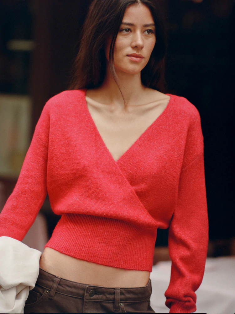 Woman wearing a red wrap-style cropped knit sweater with ribbed waistband and brown trousers against a dark background