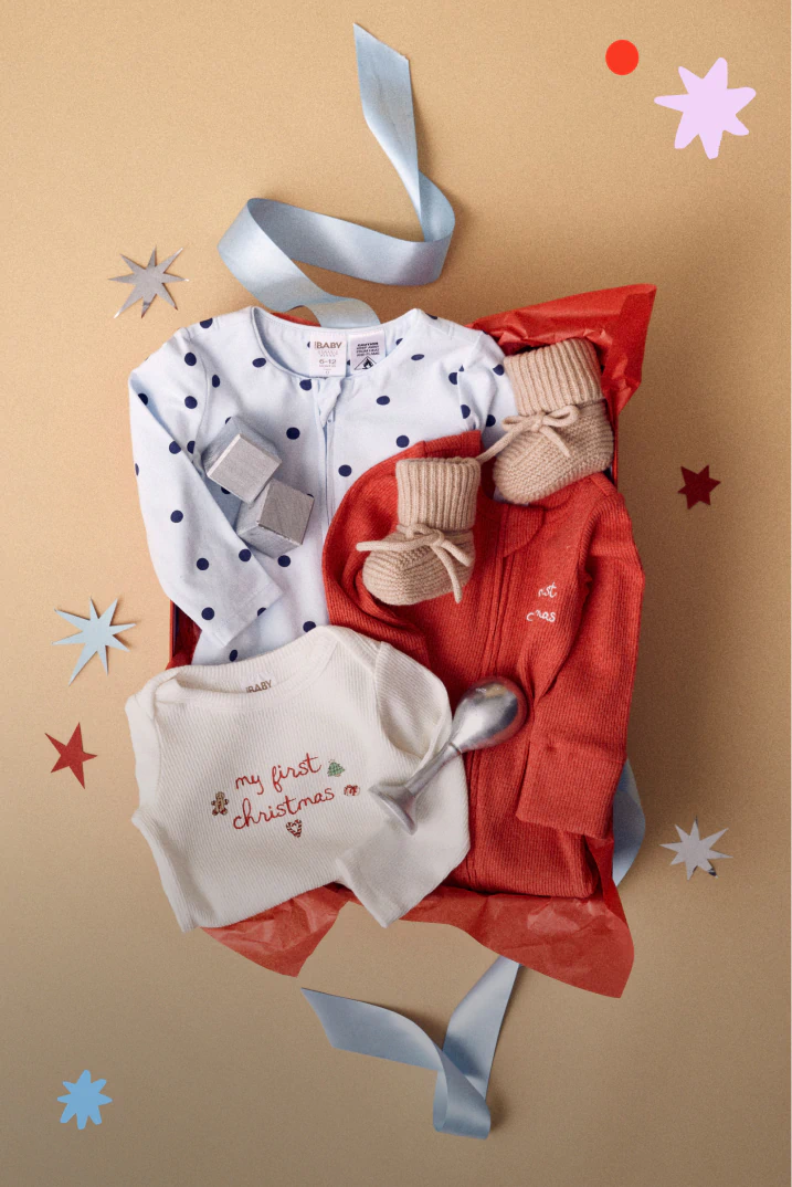 Baby's first Christmas gift set displayed flat lay: white polka dot bodysuit, cream "my first christmas" top, red cardigan, and beige socks on tan background with decorative ribbons and stars