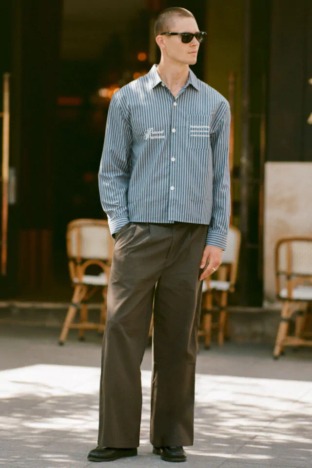 Man wearing light blue striped long-sleeve shirt and olive green pants, styled with dark sunglasses and black shoes
