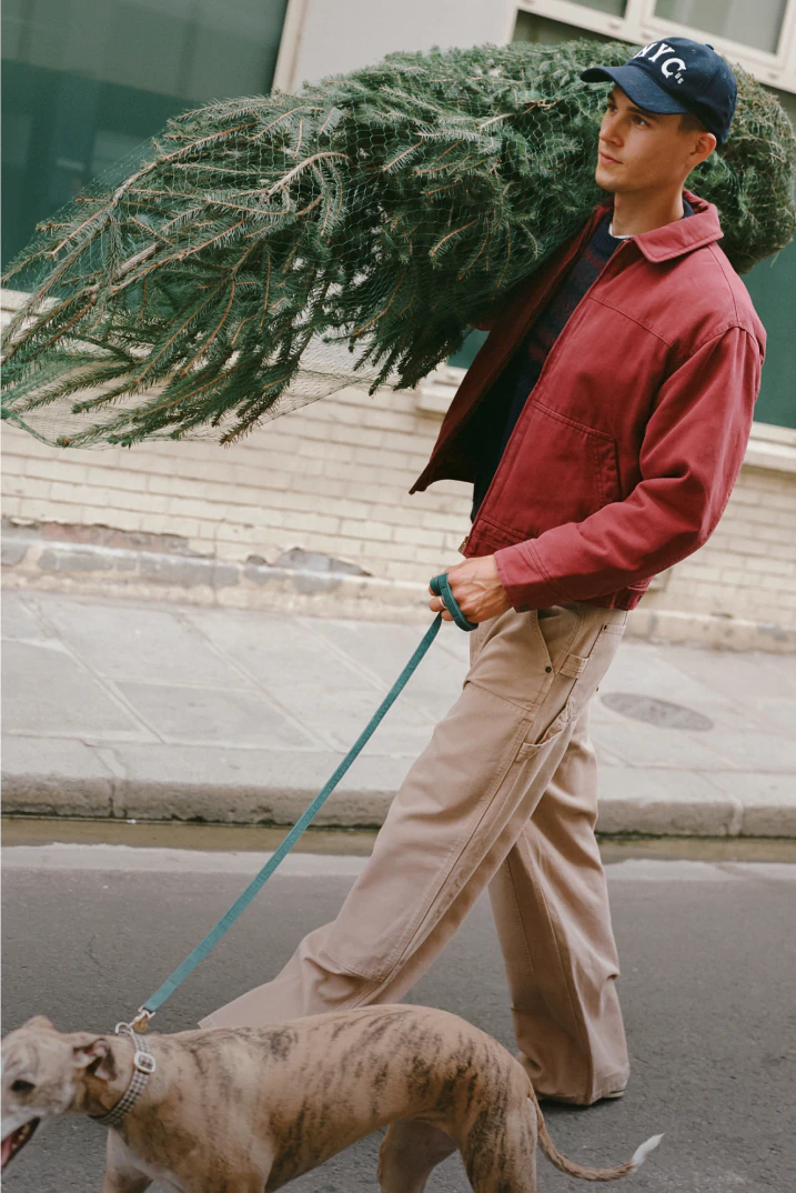 Man wearing red jacket, beige cargo pants, and navy cap carrying evergreen wreath while walking dog on city street