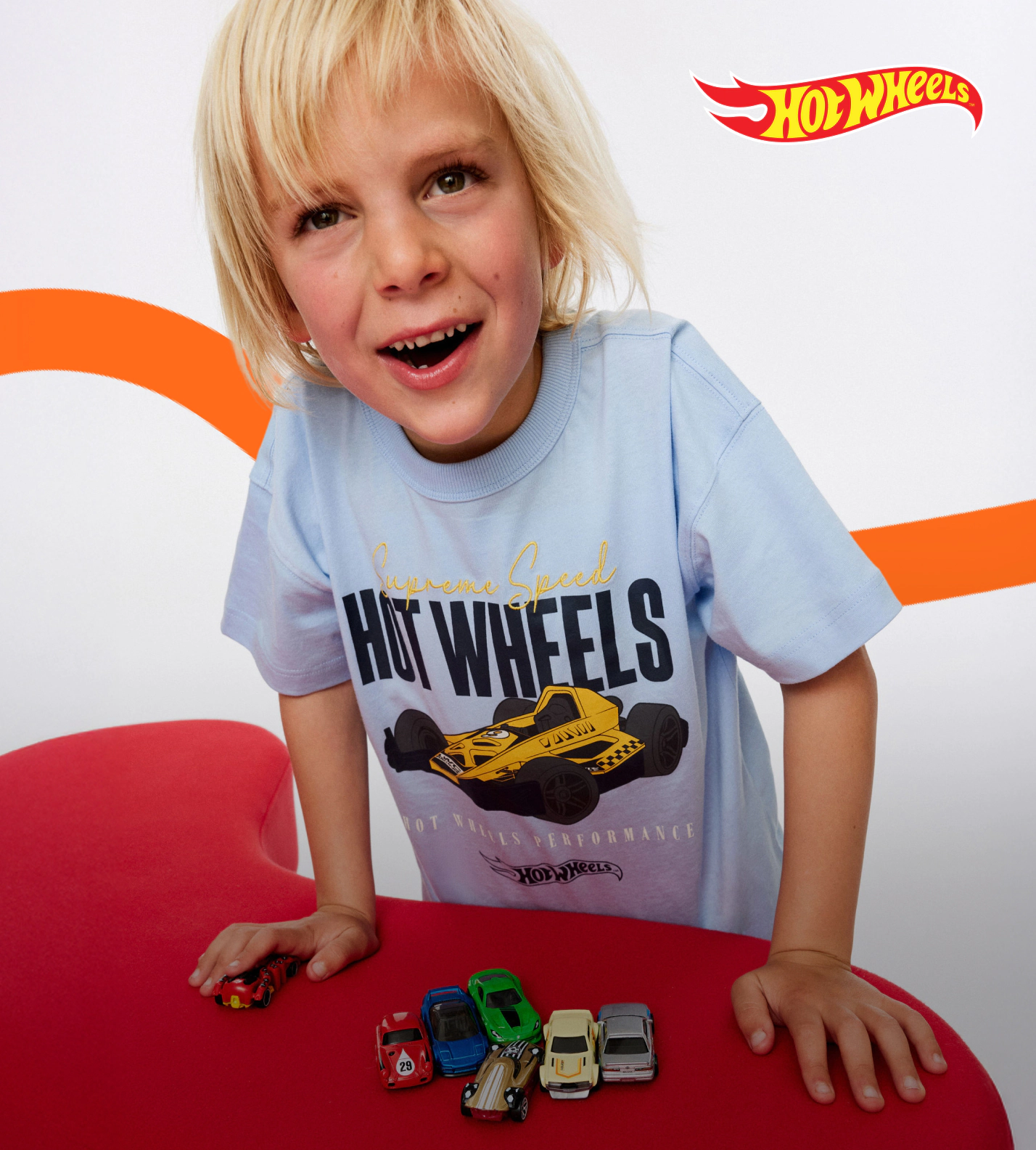 Young child with blonde hair wearing light blue Hot Wheels t-shirt, smiling at collection of miniature toy cars on red surface