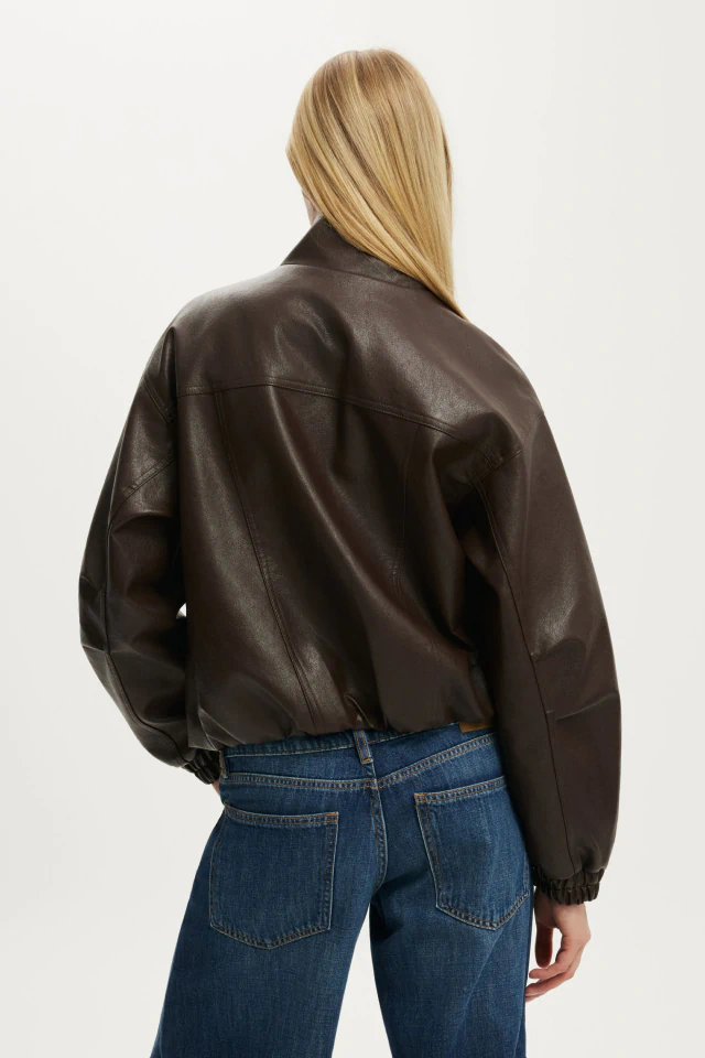Woman wearing a cropped dark brown leather jacket and blue jeans, viewed from behind against a white background