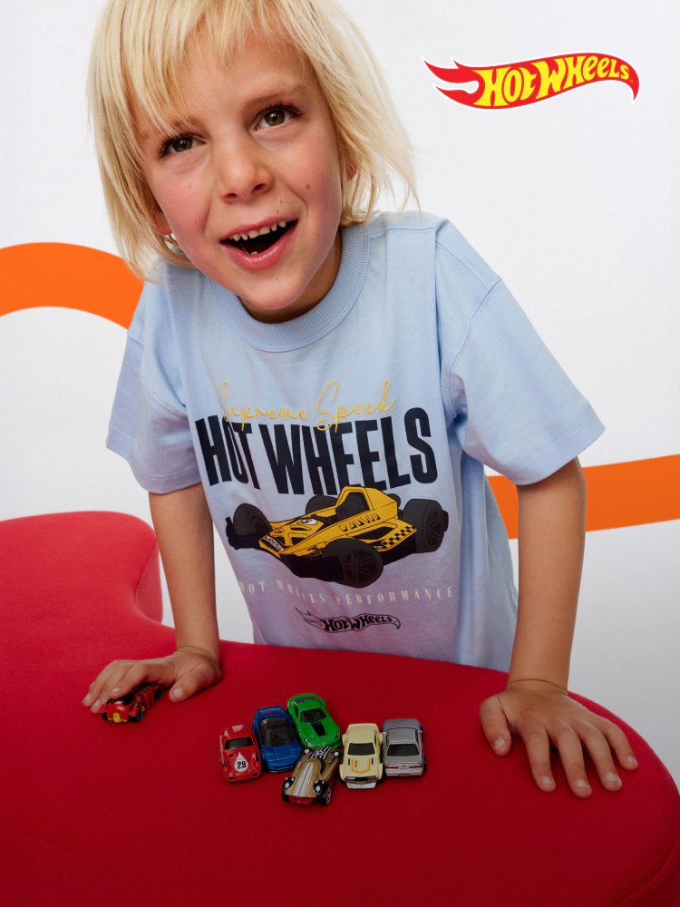 Young child wearing light blue Hot Wheels t-shirt with yellow race car graphic, displaying miniature Hot Wheels vehicles on red surface with Hot Wheels logo above