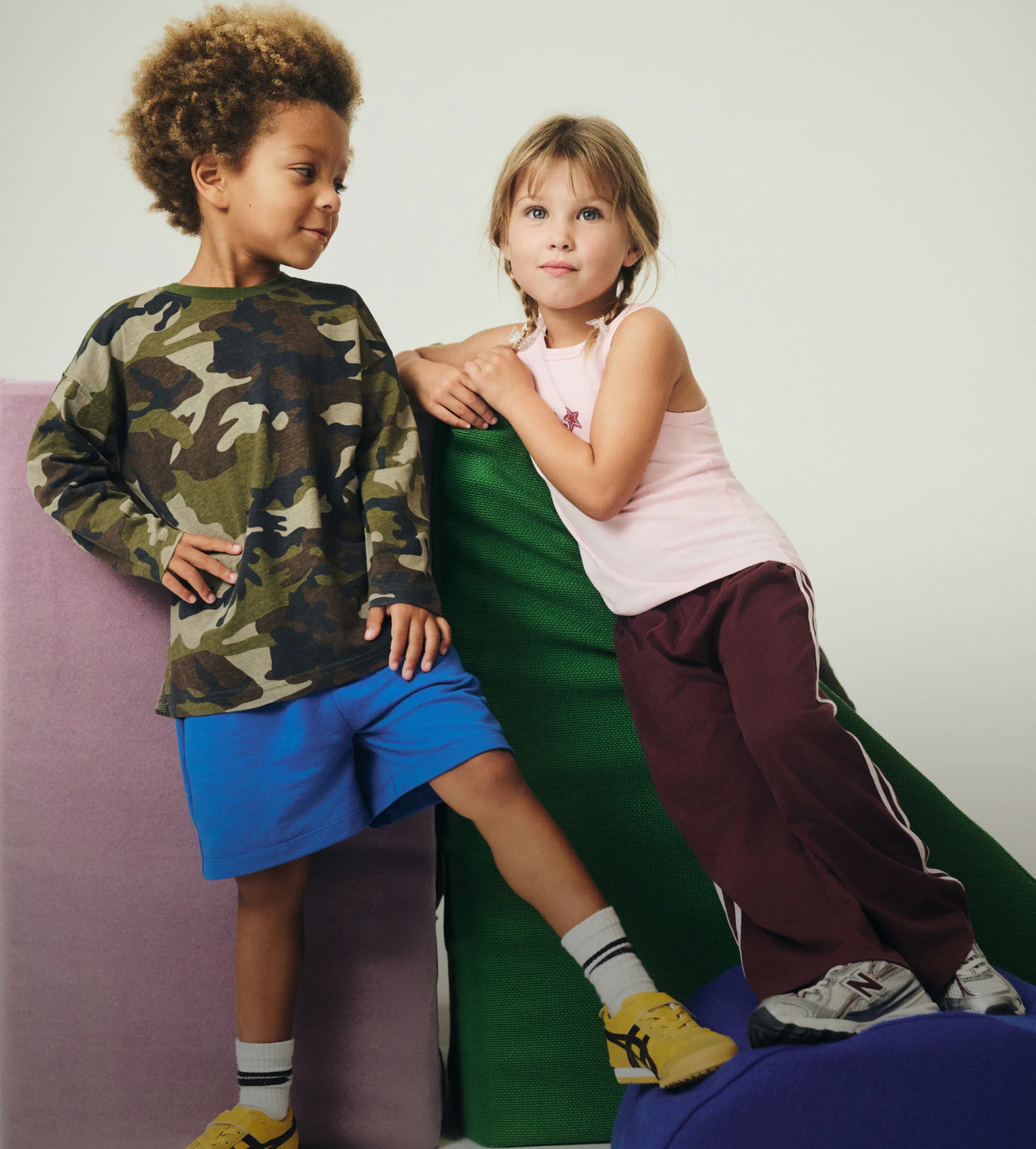 Two children posing together against a neutral background wearing colorful casual clothing including camouflage print and knit pieces