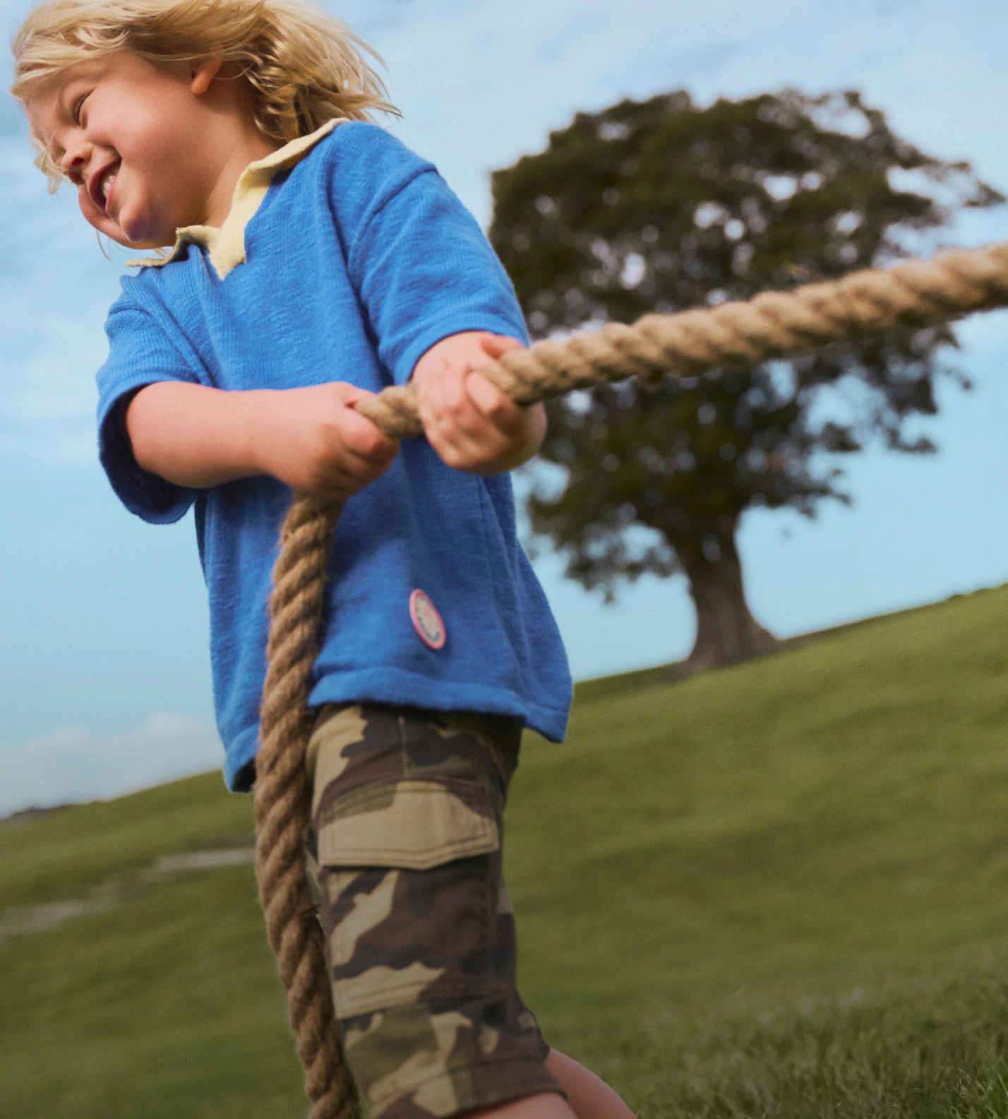 Young child in blue t-shirt pulling a rope outdoors with tree and green field in background, pixelated parrot graphic overlaid