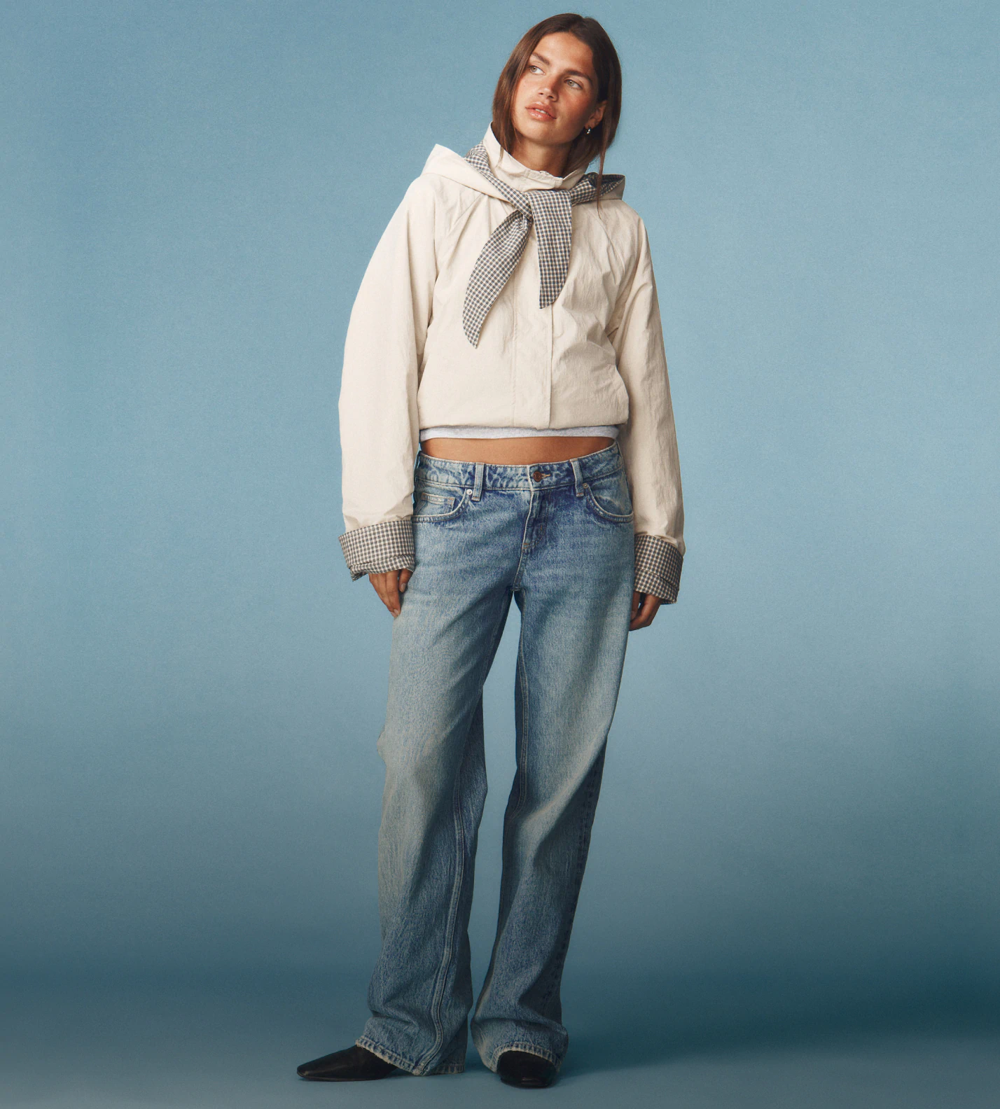 Woman wearing navy three-quarter sleeve raglan shirt over white long-sleeve top, light-wash jeans, black belt, and black shoulder bag against blue background
