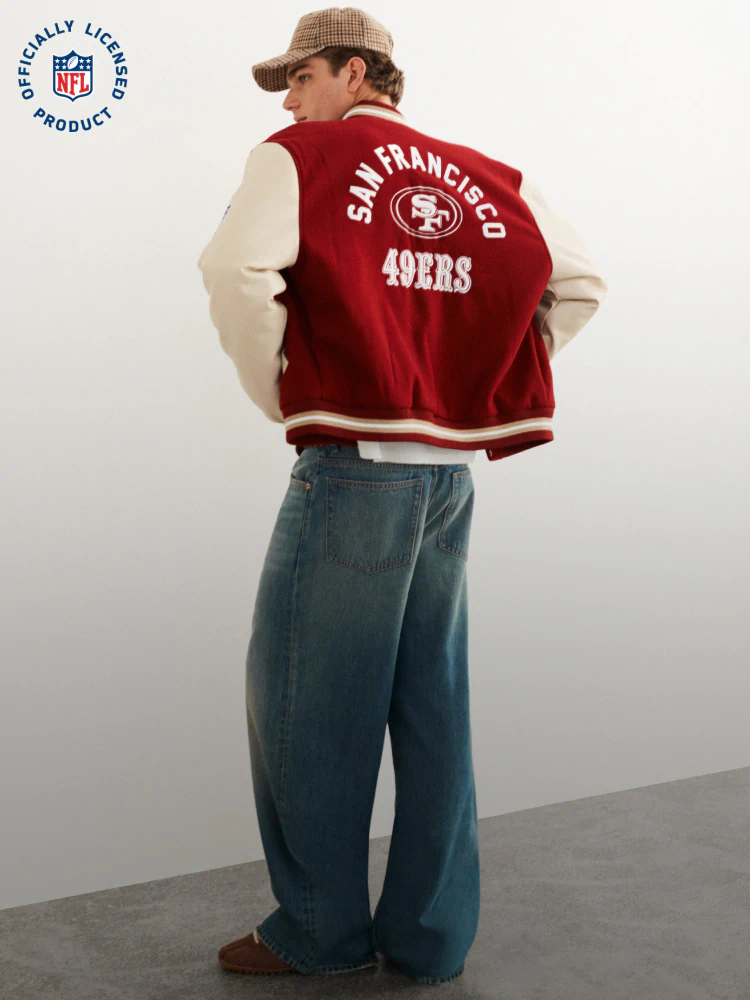 Man wearing San Francisco 49ers red and cream varsity jacket over cream long-sleeve shirt, blue jeans, and tan cap, shown from behind