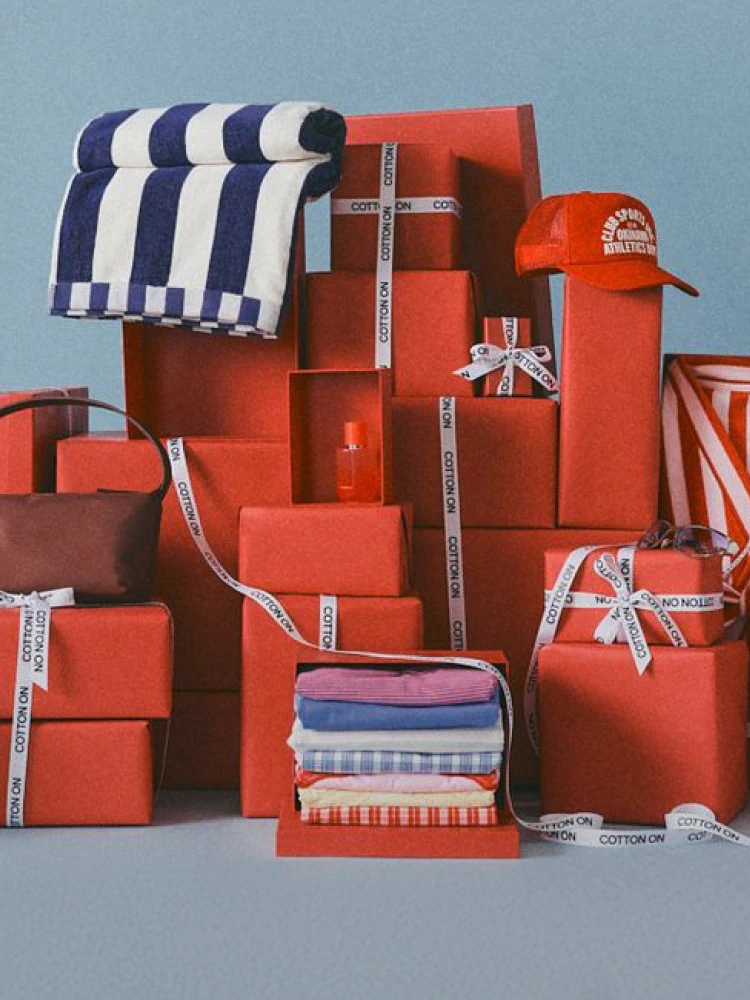 Cotton On red gift boxes stacked in pyramid arrangement with navy striped towel, folded clothing, orange cap, and "Cotton On" branded ribbons against blue background