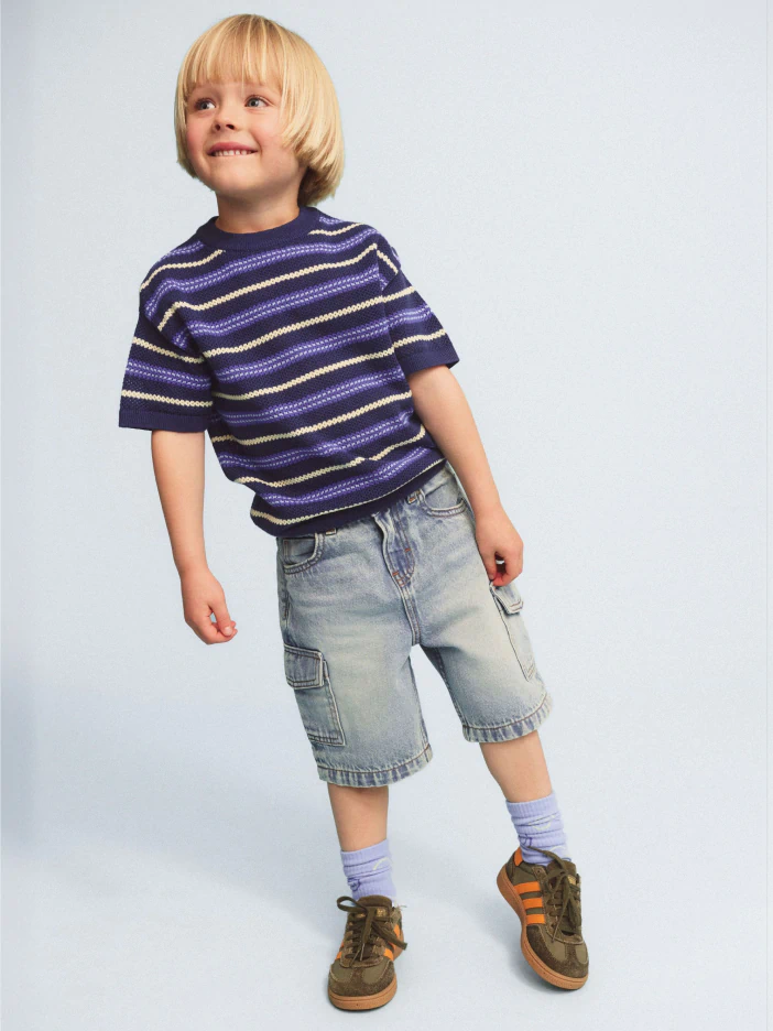 Young child with blonde bob hair wearing a purple striped short-sleeve t-shirt, light blue cargo shorts, and brown sneakers with orange accents