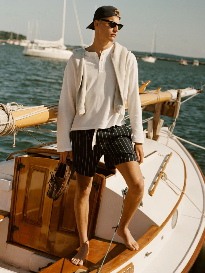 Man wearing cream linen shirt and black pinstriped shorts standing on sailboat with water and sailing vessels in background