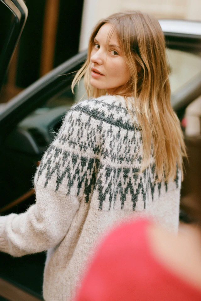 Woman with long reddish-brown hair wearing a cream and green patterned knit sweater, photographed outdoors