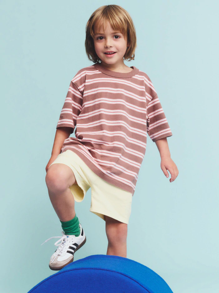 Young child wearing a pink and white striped long-sleeve shirt and cream shorts, standing on a blue ball against a light blue background