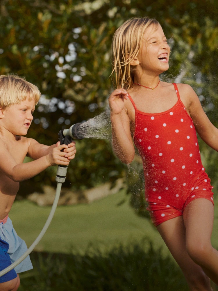 Two children playing with a garden hose outdoors. Boy in blue shorts, girl in red polka dot swimsuit. Pixelated parrot illustration overlaid