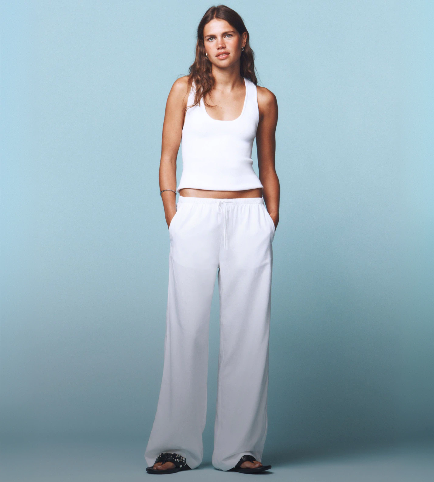 Woman wearing white sleeveless crop top and white wide-leg pants with black slide sandals against blue background