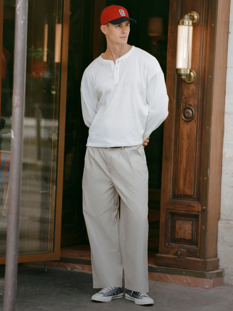 Man wearing red baseball cap, white long-sleeve henley shirt, and beige pants standing in front of wooden doors