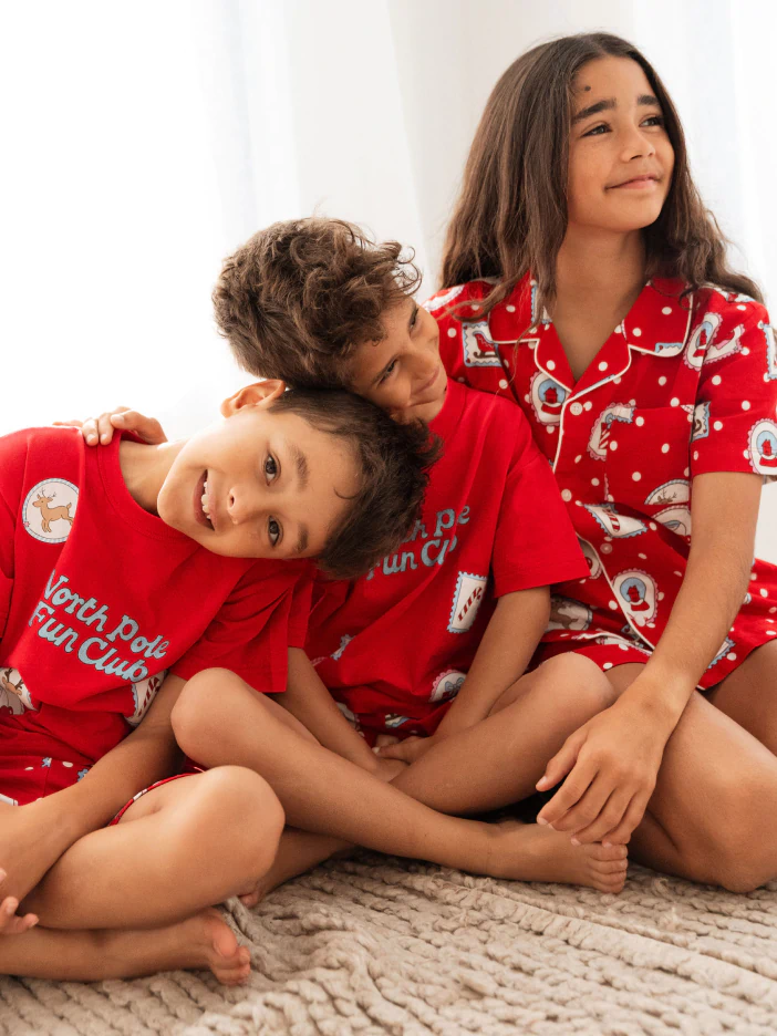 Three children wearing red festive pajamas with reindeer and snowflake patterns, sitting together on a beige textured surface