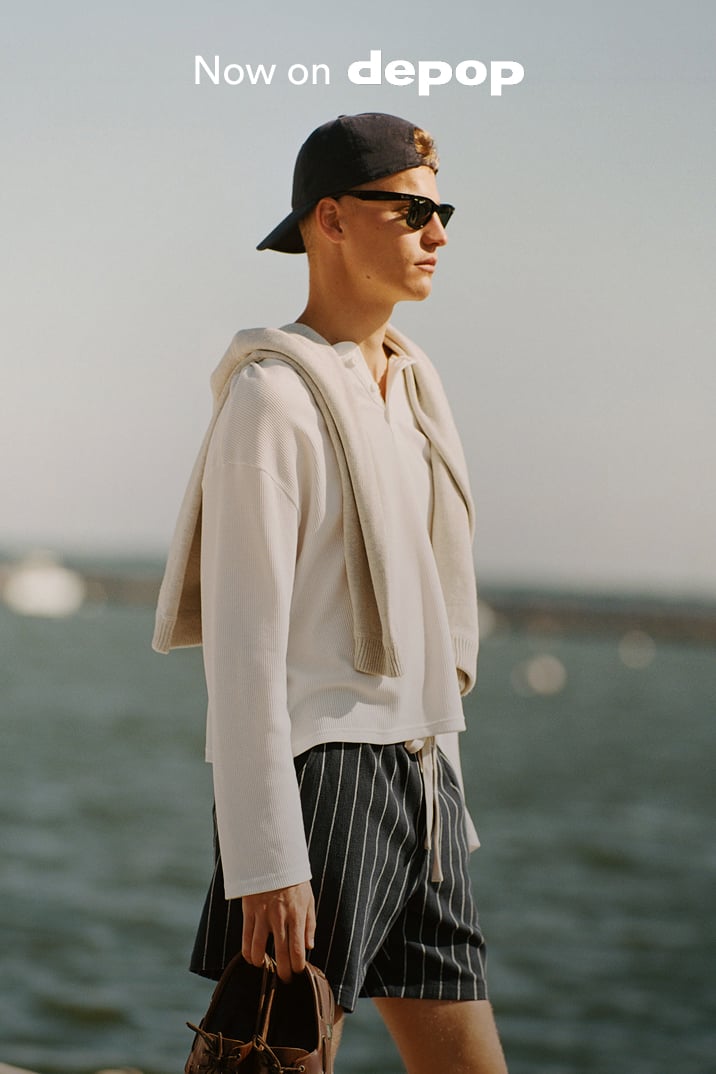 Man wearing cream hoodie and black pinstriped shorts with brown bag, standing by water. Text reads "Now on depop