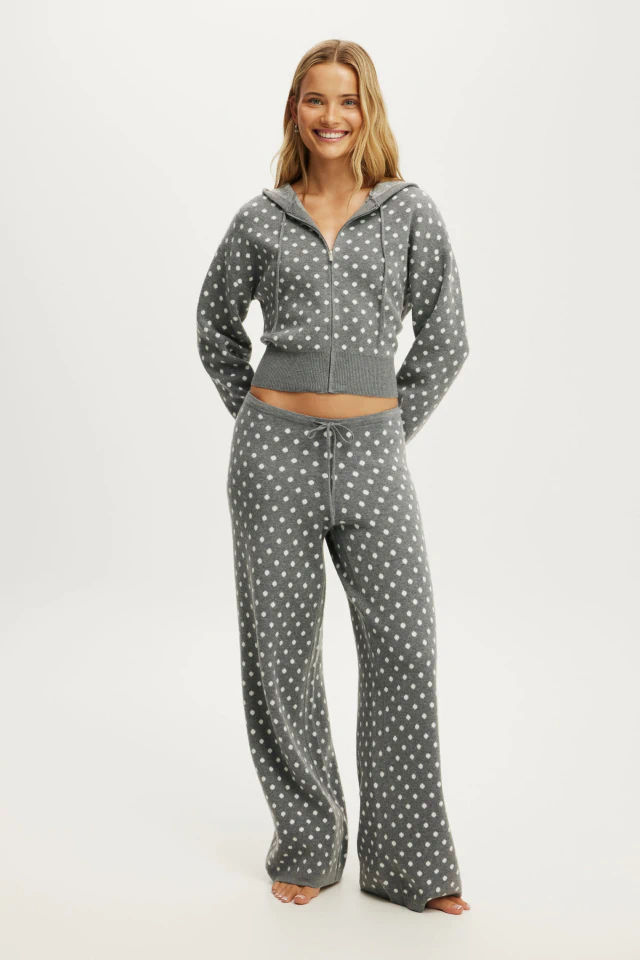 Woman wearing gray polka dot cropped zip-up hoodie and matching wide-leg pajama pants, smiling at camera against white background