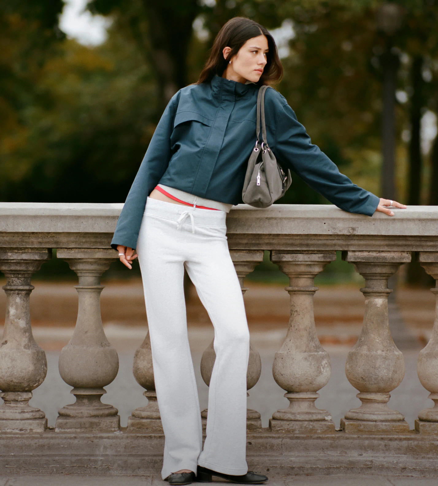 Woman wearing teal blue cropped jacket, white flared pants with red waistband, and gray shoulder bag, leaning against stone railing outdoors