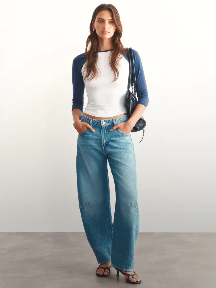 Woman wearing navy raglan sleeve top, white cropped tee, and blue jeans with black shoulder bag and sandals against white background