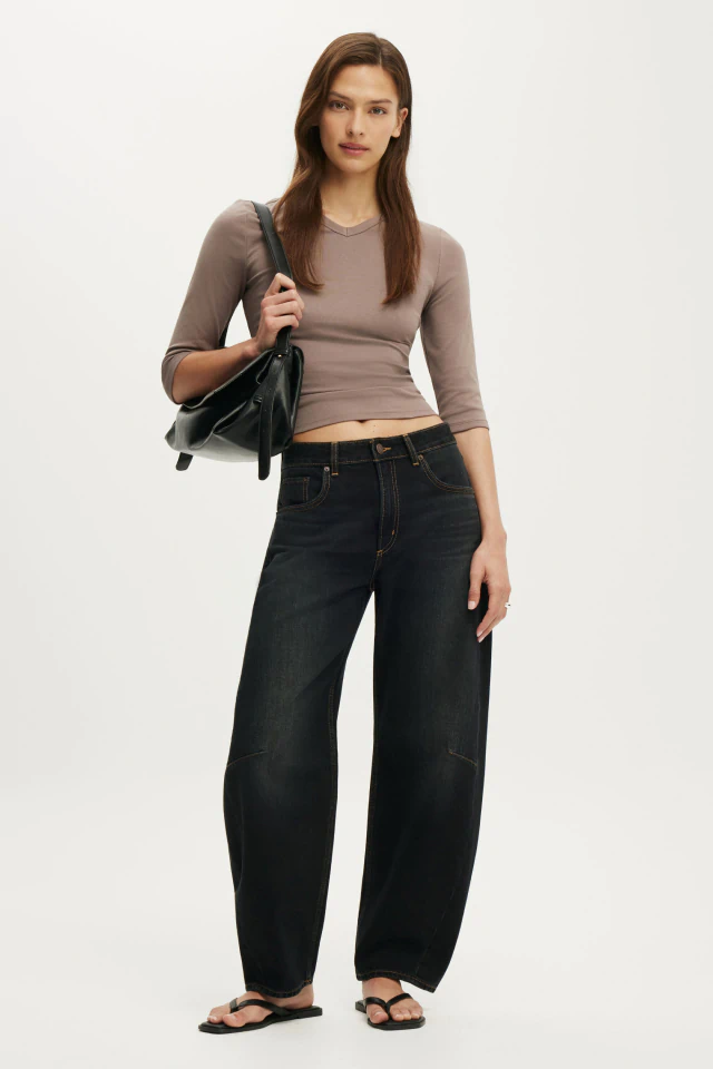 Woman wearing taupe long-sleeve crop top and dark wash wide-leg jeans, carrying black shoulder bag, standing against white background