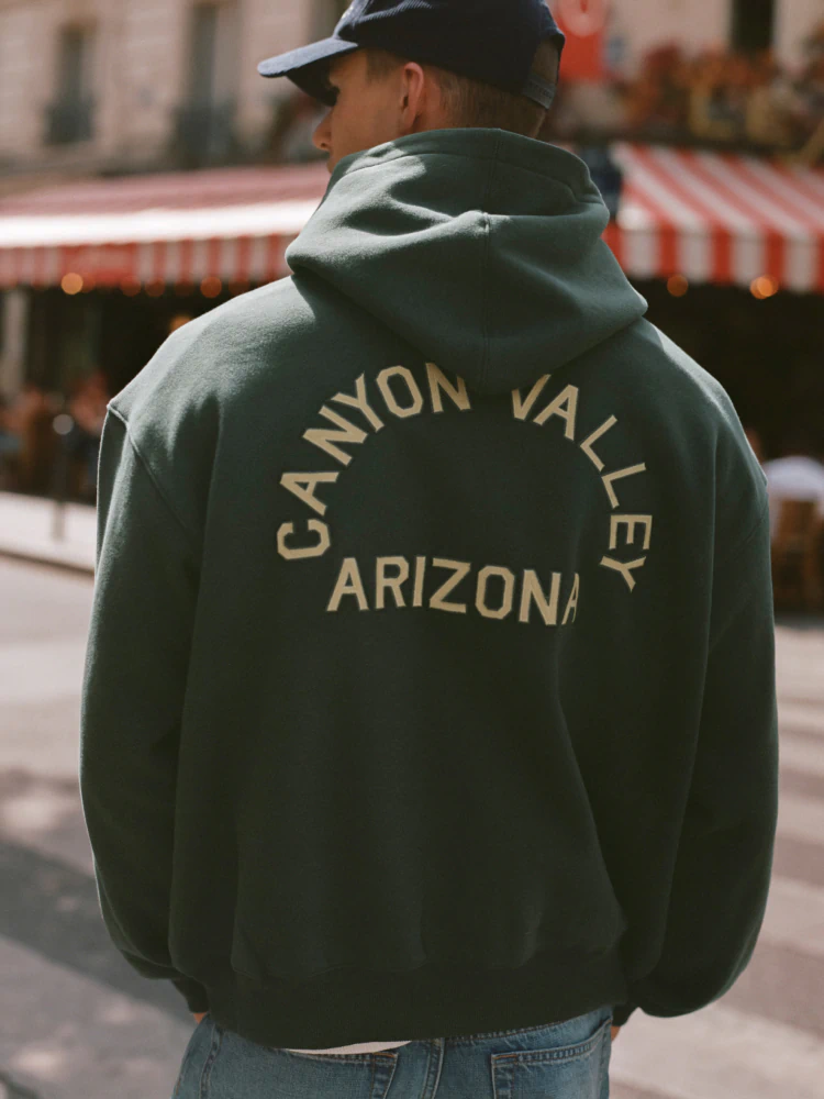 Person wearing dark green hoodie with "Canyon Valley Arizona" text in tan lettering, navy cap, viewed from behind on street with striped awning background