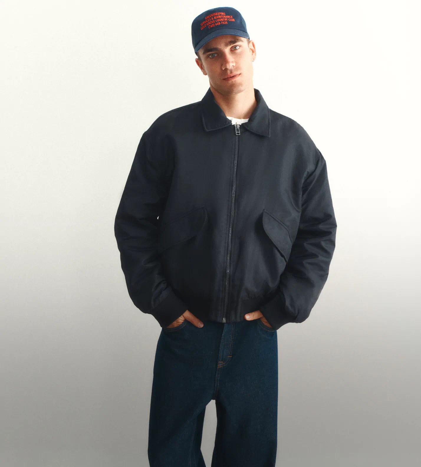 Man wearing dark navy zip-up jacket and dark blue jeans with navy cap featuring red text