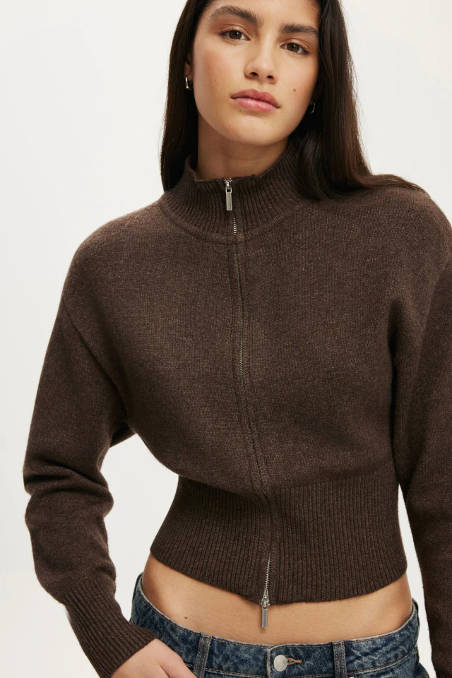 Woman wearing a brown ribbed zip-up cropped cardigan over a denim waistband against a white background